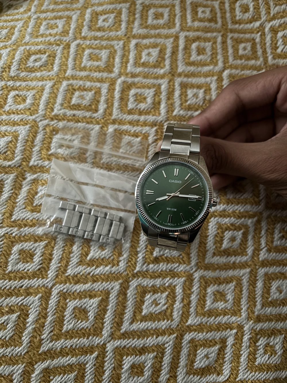 A steel watch with a green dial held in one hand, its bracelet lying loose in a bag beside it